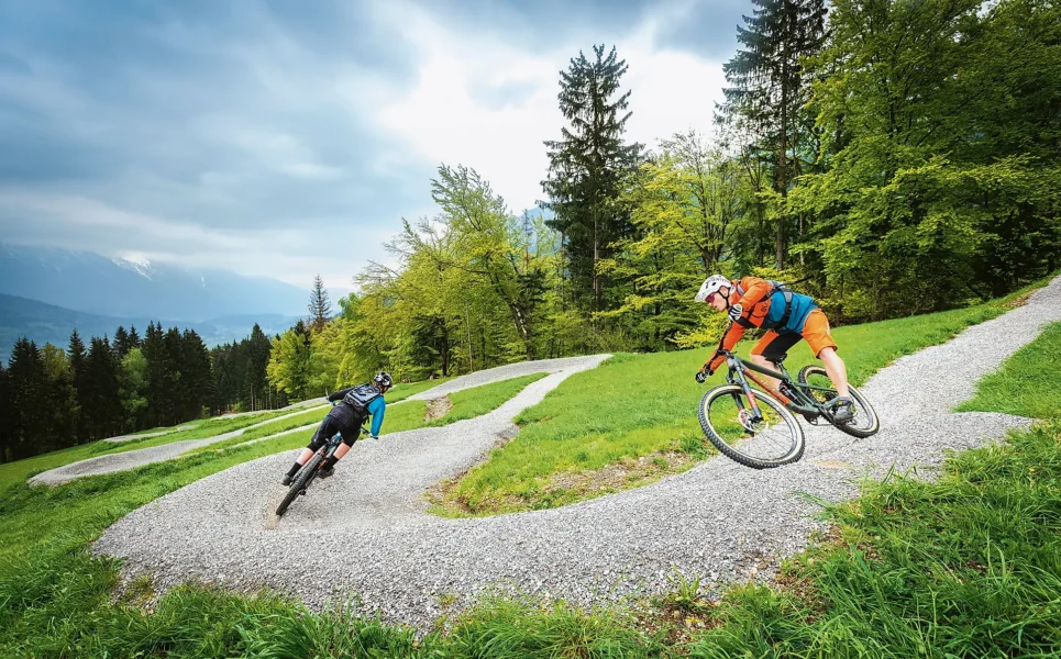 Twee personen op een mountainbike die over een grindtrail de berg af fietsen. Op de achtergrond dennenbomen.
