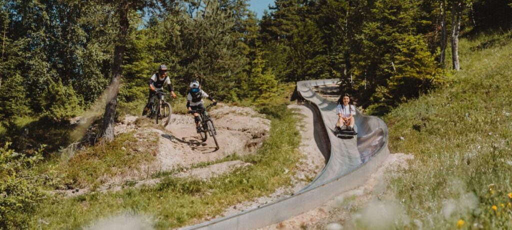 Vrouw rodelt van zomerrodelbaan "Wildcat" op de Katzenkopf in Leutasch met ernaast twee mensen op mountainbike die over een bergpad in het naastgelegen bikepark fietsen.