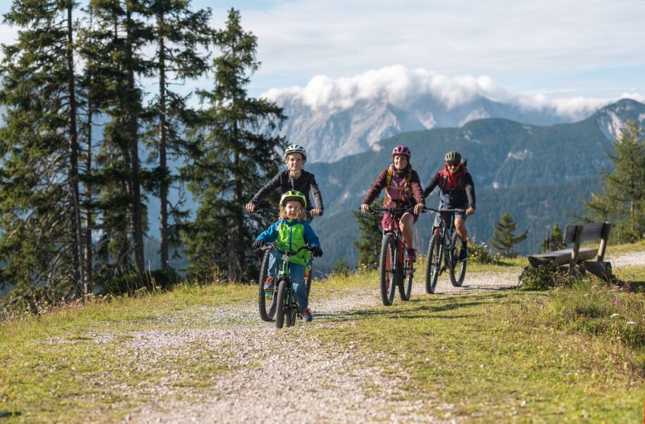 Gezin fietst op mountainbikes over een breed bergpad met bos en bergen op de achtergrond in de omgeving van Seefeld en Leutaschtal.